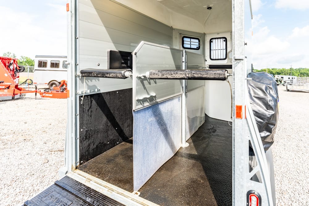 Interior of a horse trailer showing a sliding divider and central stall barrier, with rubberized floor and light metal walls outside on a gravel lot
