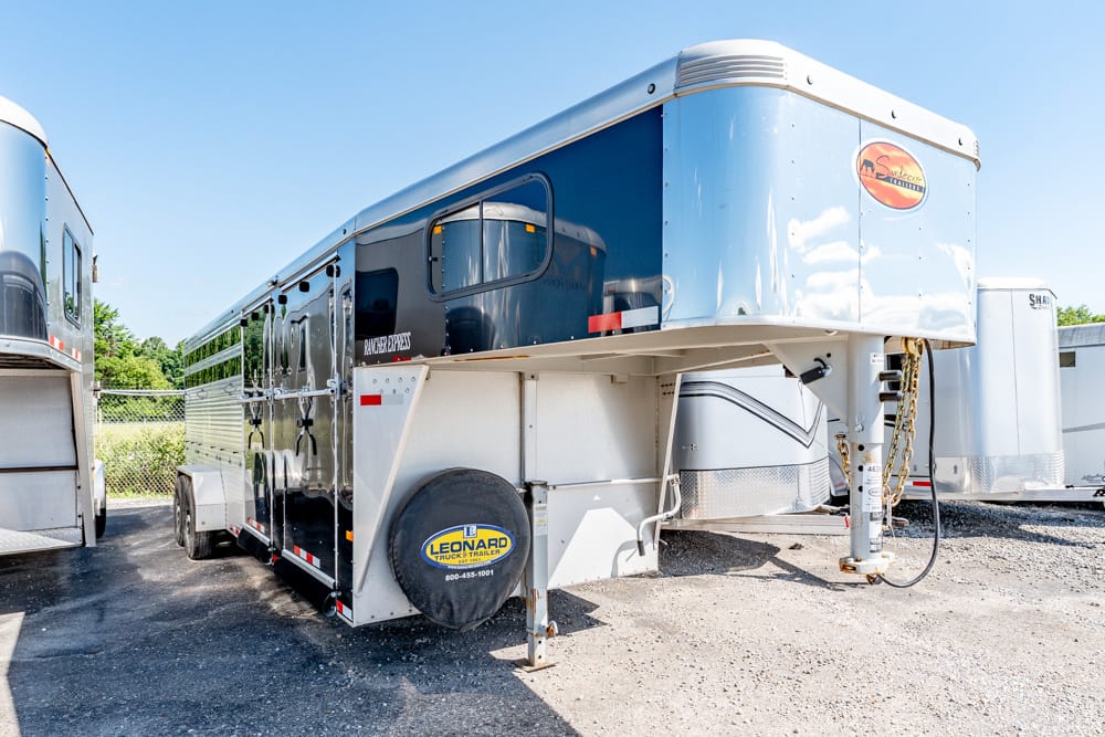 Large gooseneck trailer parked in a lot, with a spare tire cover and blue-and-gray exterior under a clear sky
