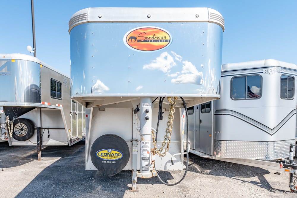 Blue gooseneck horse trailer with Sundowner Trailers emblem, parked between other aluminum trailers on a gravel lot, showing hitch, chains, and spare tire.