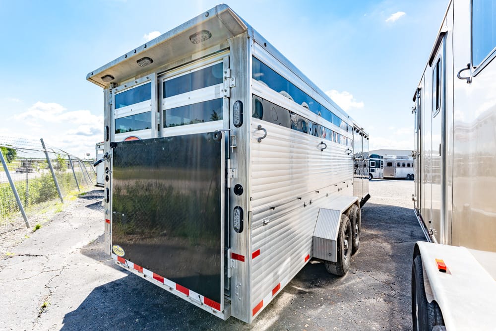 Aluminum horse trailer with a closed rear ramp, parked in a lot beside another trailer on a sunny day.