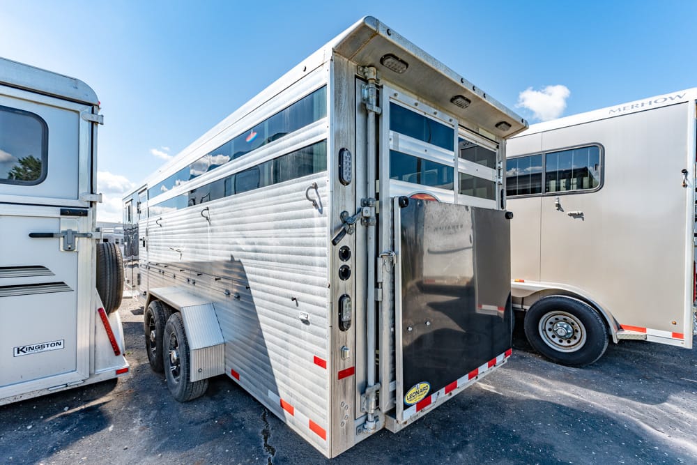 Aluminum livestock trailer with its rear ramp or gate closed, parked between other horse trailers on a sunny day