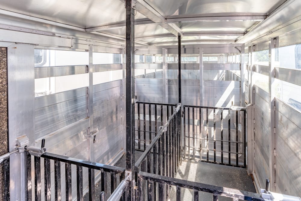 Interior of a metal livestock trailer with partitioned stalls and black gates, sunlight streaming through side vents