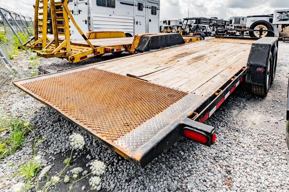 Flatbed trailer with a rusted perforated ramp, parked on a gravel lot with other trailers in the background.