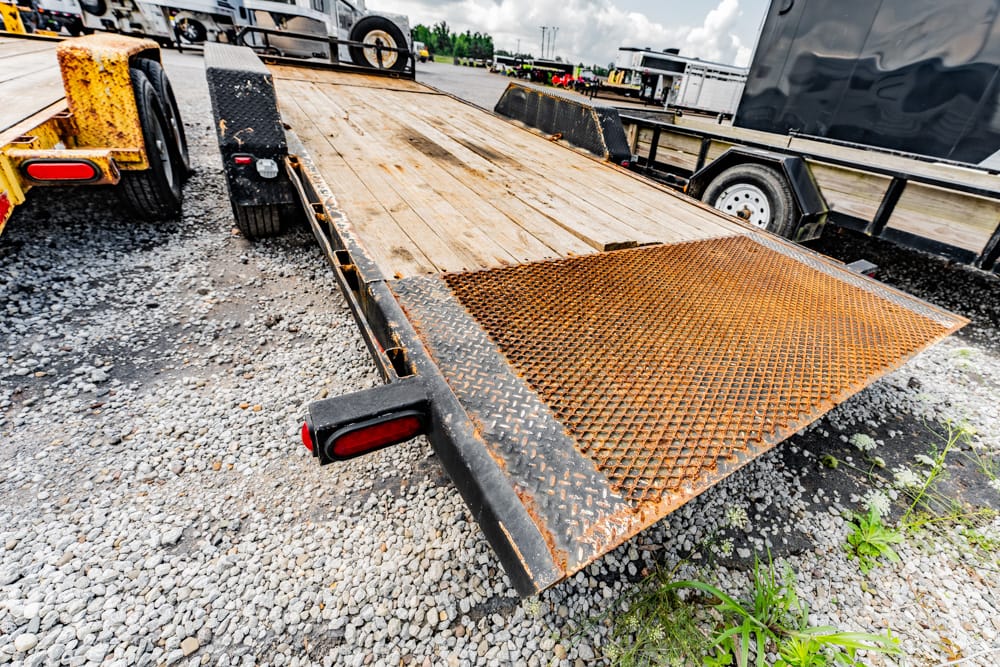Rusty metal car trailer with a folded ramp on a gravel lot, other trailers in the background.