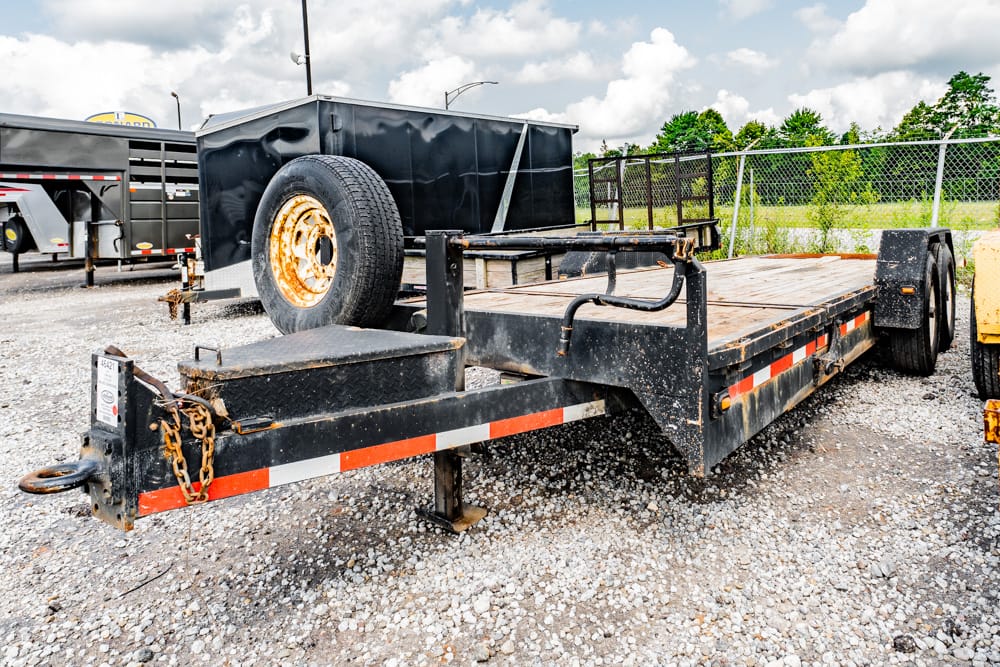 Flatbed trailer with wooden deck and spare tire on the front, parked on a gravel lot with chains and safety features visible on the hitch.