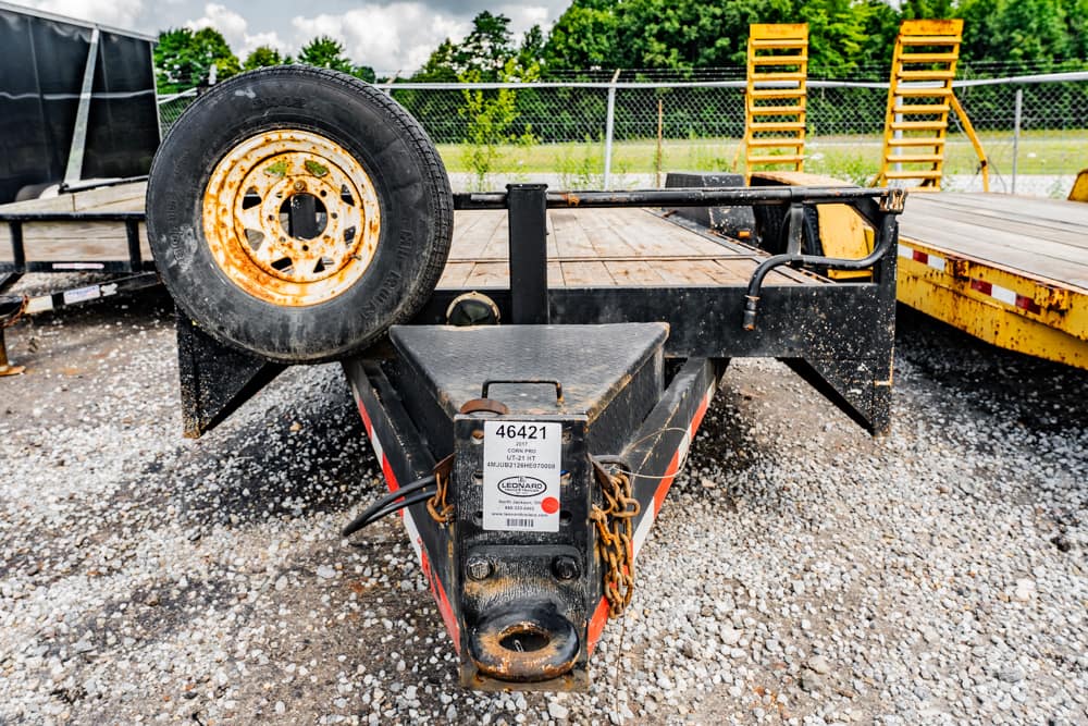 Front view of a utility trailer hitch with a mounted spare tire on the left, rusty chains, and a gravel yard background.