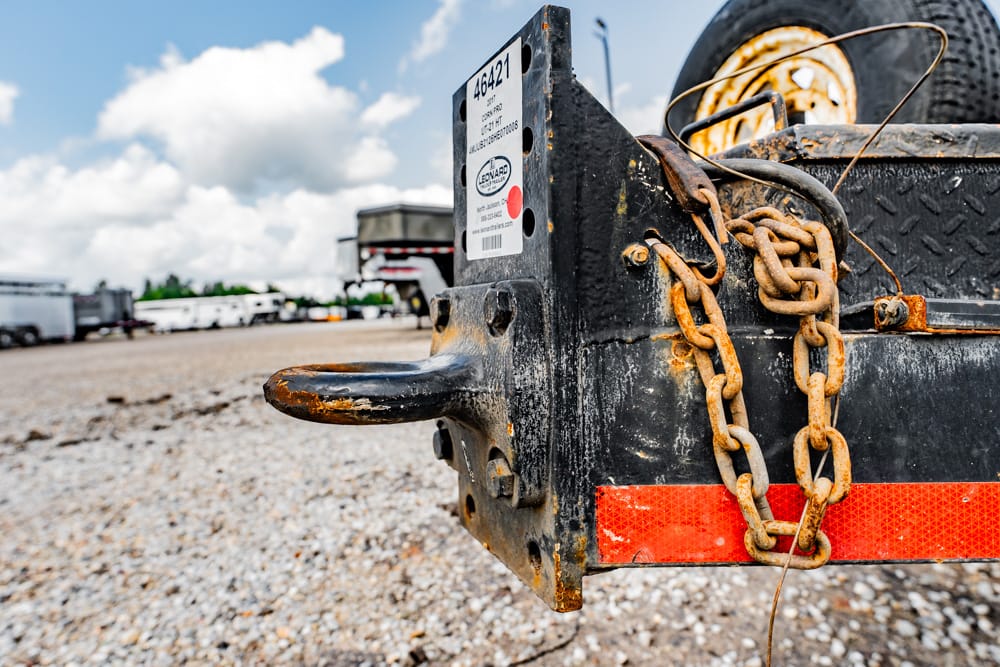 Close-up of a rusted trailer hitch with heavy chains on a black trailer in a gravel lot outside an industrial yard.