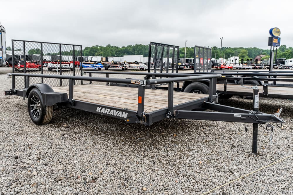 Karavan flatbed trailer with a wooden deck and metal railing on a gravel lot, parked among other trailers.