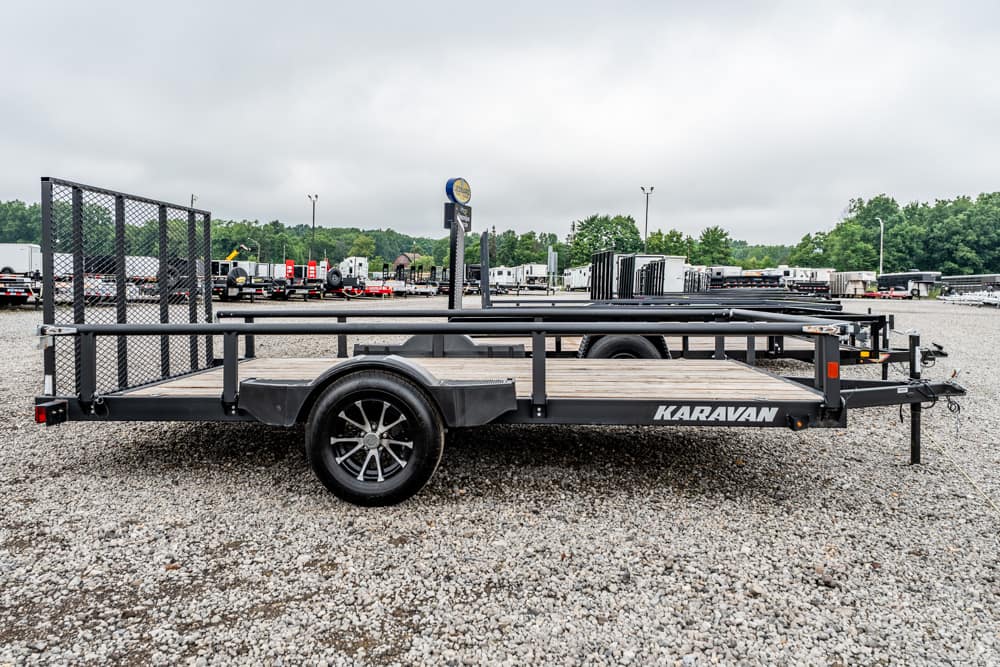 Black Karavan flatbed trailer with a wooden deck and mesh rear gate parked on a gravel lot at a trailer yard. Note: concise description of the trailer and setting.