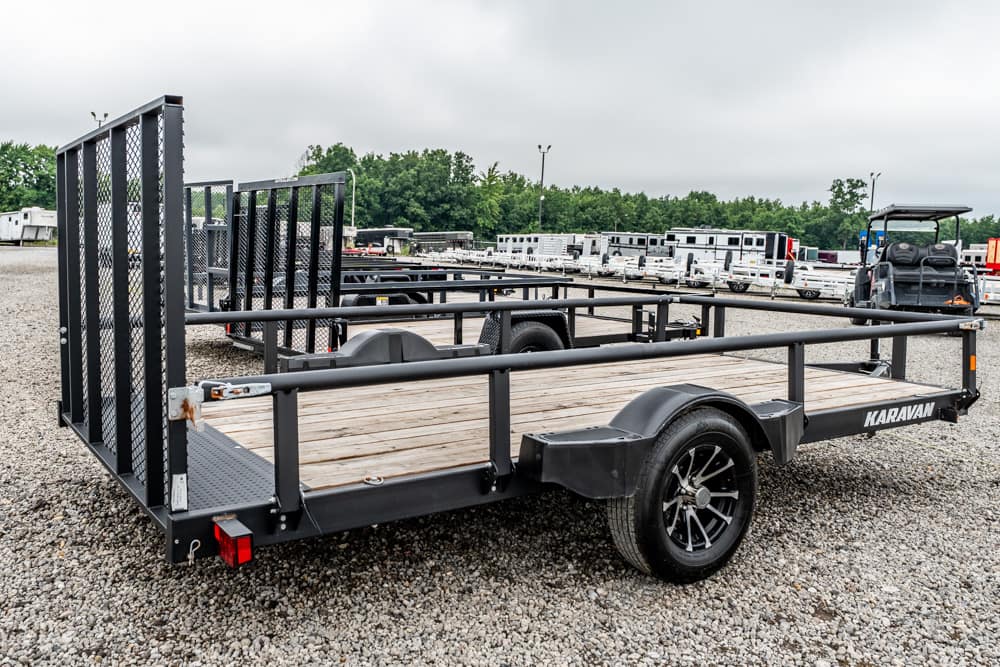 Black flatbed utility trailer with wooden deck and high side rails, labeled 'KARAVAN', parked on gravel at a trailer lot