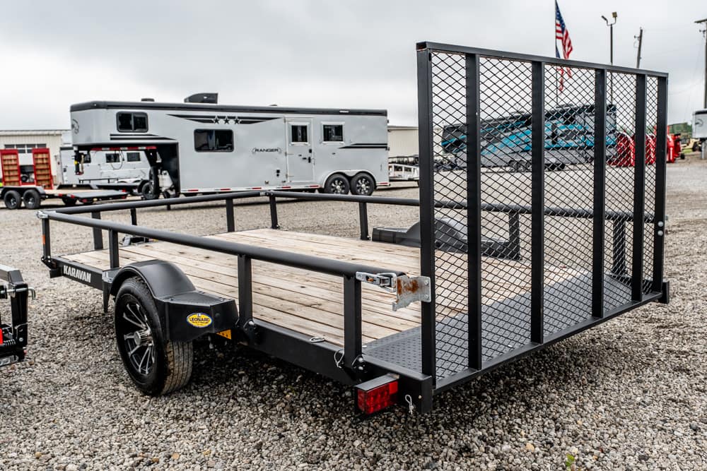 Flatbed utility trailer with wooden deck and a tall mesh gate on a gravel lot; a silver horse trailer is visible in the background.