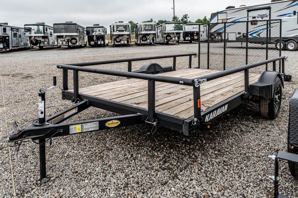 Flatbed trailer with a wooden deck and side rails in a gravel lot, with parked trailers and RVs in the background.