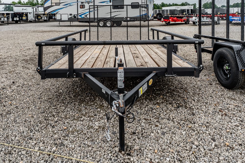 Front view of a flatbed utility trailer with a wooden deck and black metal frame on a gravel lot, hitch and jack in foreground