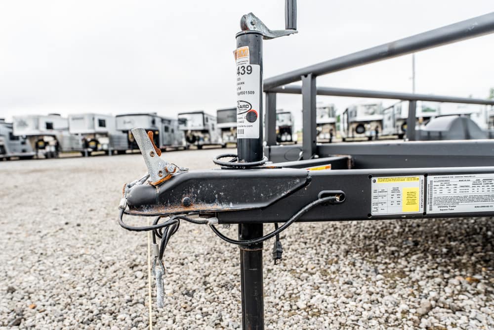 Close-up of a trailer hitch and jack on a black flatbed trailer with a gravel lot and rows of trailers in the background