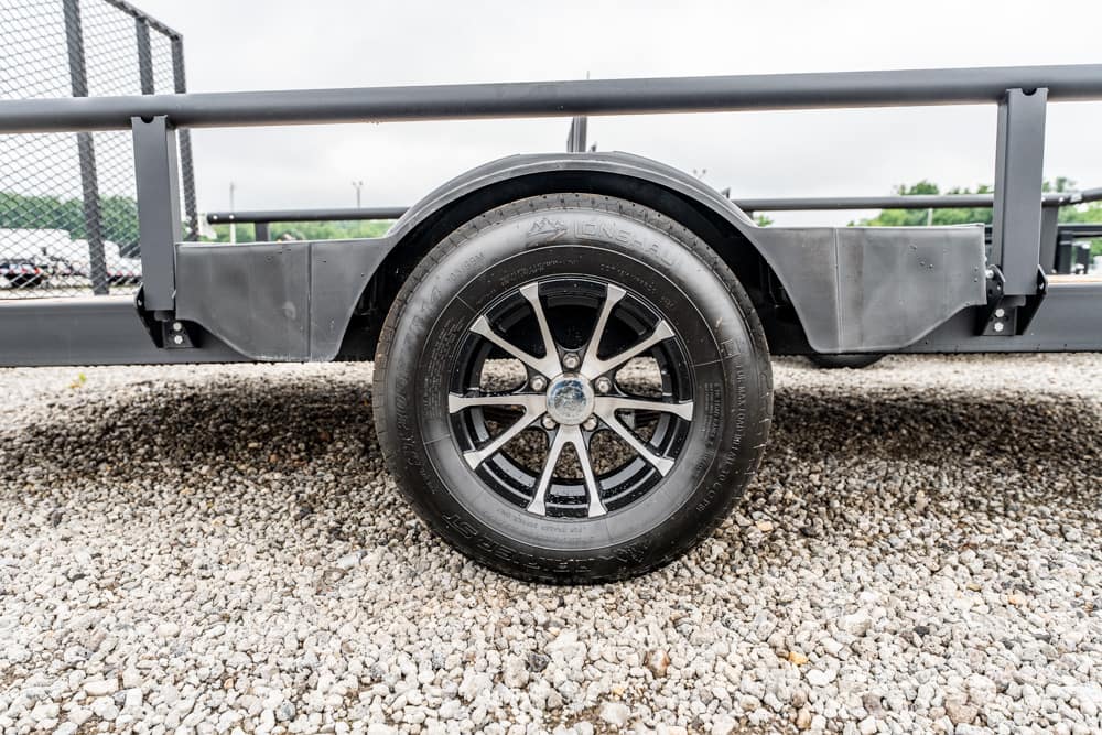 Black trailer wheel with chrome spokes mounted on a gray trailer frame over a gravel lot.
