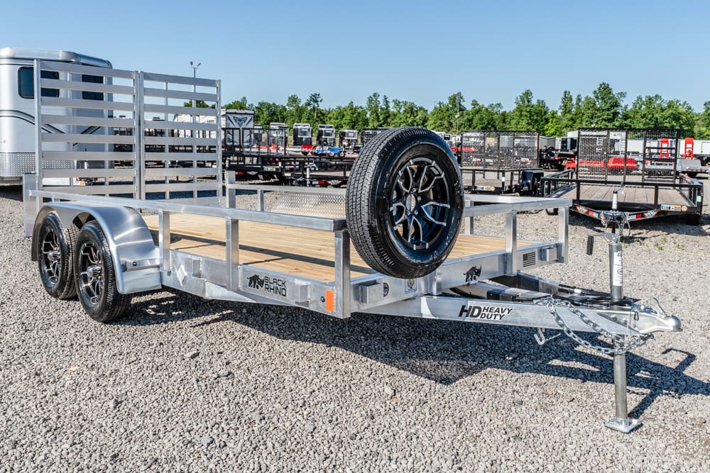 Aluminum utility trailer with a wooden deck, spare tire mounted on the front, and dual-axle wheels in a gravel lot.
