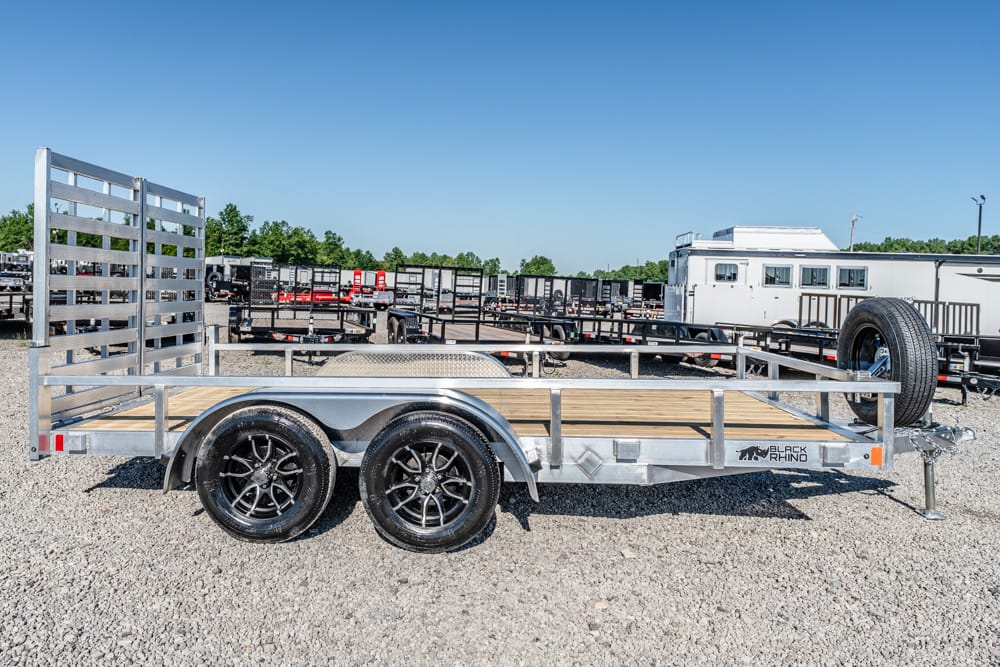 Silver double-axle utility trailer with a wooden deck and ramp gate, parked on a gravel lot, spare tire mounted at the front.