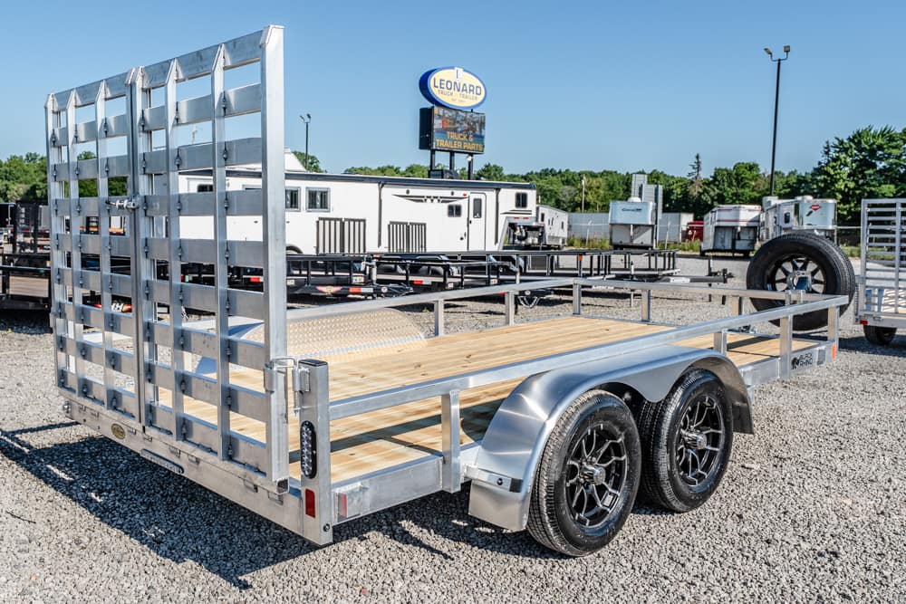 Aluminum flatbed trailer with a wooden deck and dual axles parked on a gravel lot at a trailer store. A 'Leonard' sign is visible in the background.