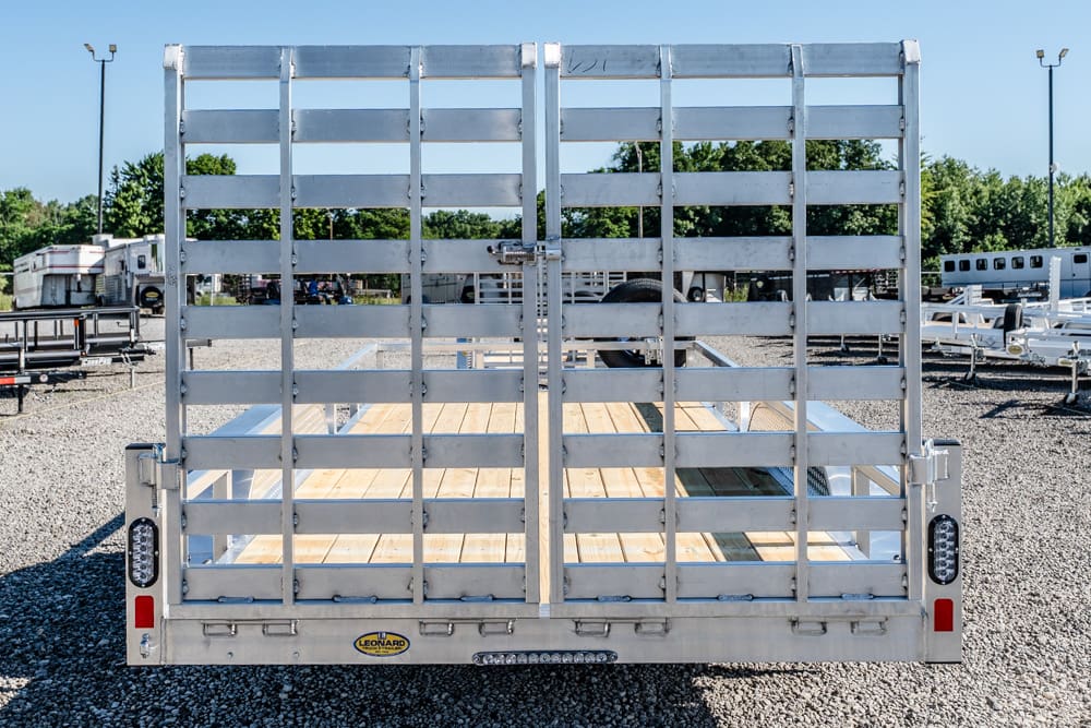 Rear view of a silver utility trailer with a wooden bed and slatted vertical gate, parked in a gravel lot with other trailers in the background.