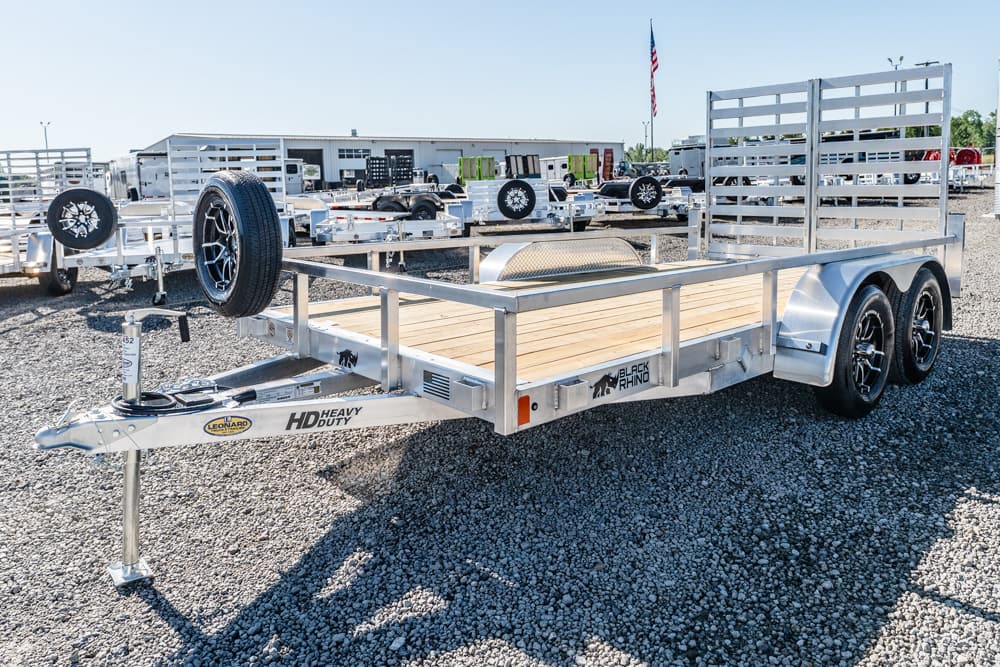 Aluminum flatbed utility trailer with a wooden deck and dual axles parked on a gravel lot, branding visible on the frame.