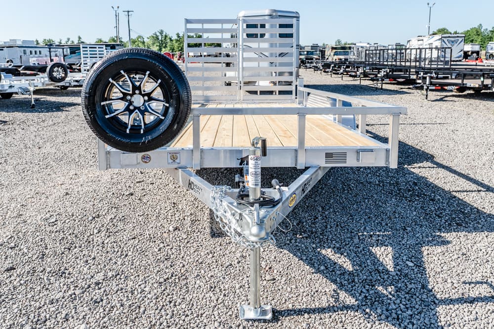Front view of a flatbed utility trailer with a mounted spare tire in a gravel lot.