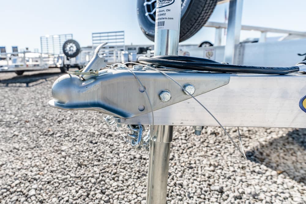 Close-up of a metal boat trailer hitch with bolts, safety chains, and wiring on a gravel lot.