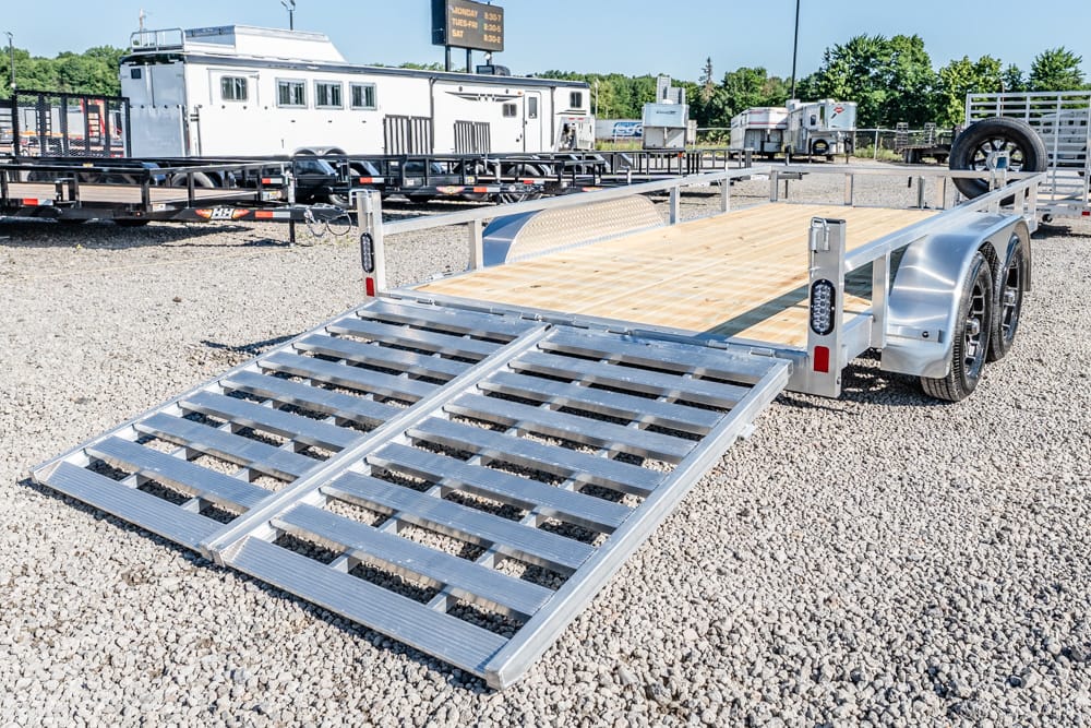 Double-axle trailers with a wooden deck and metal ramp lowered onto a gravel lot, ready for loading equipment.