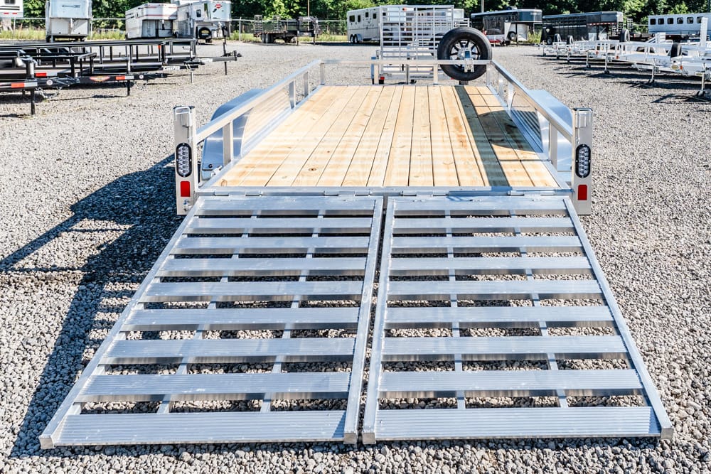 Open dual ramps on a flatbed trailer with a wooden deck, parked on a gravel lot with other trailers in the background.