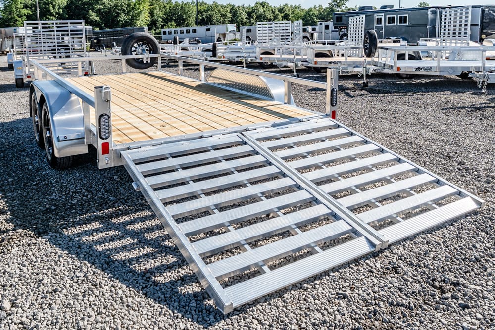 Aluminum flatbed trailer with a fold-down loading ramp extended onto a gravel lot, ready to haul vehicles or equipment.