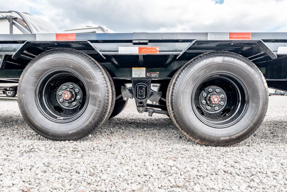 Dual-axle trailer wheels on a gravel surface, showing the tandem tires and rims under a flatbed trailer.