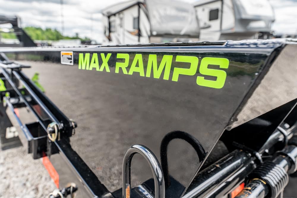 Black trailer side with bright green 'MAX RAMPS' logo in clear focus, hitch and equipment visible in foreground
