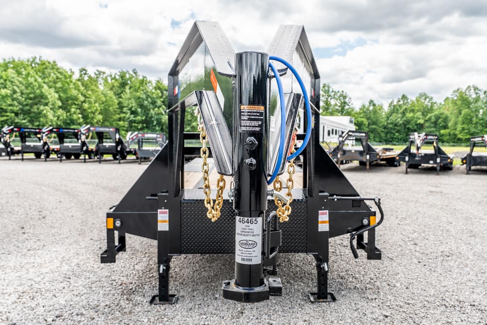Close-up of a gooseneck trailer hitch with safety chains and blue wiring in a gravel lot, with multiple trailers in the background.