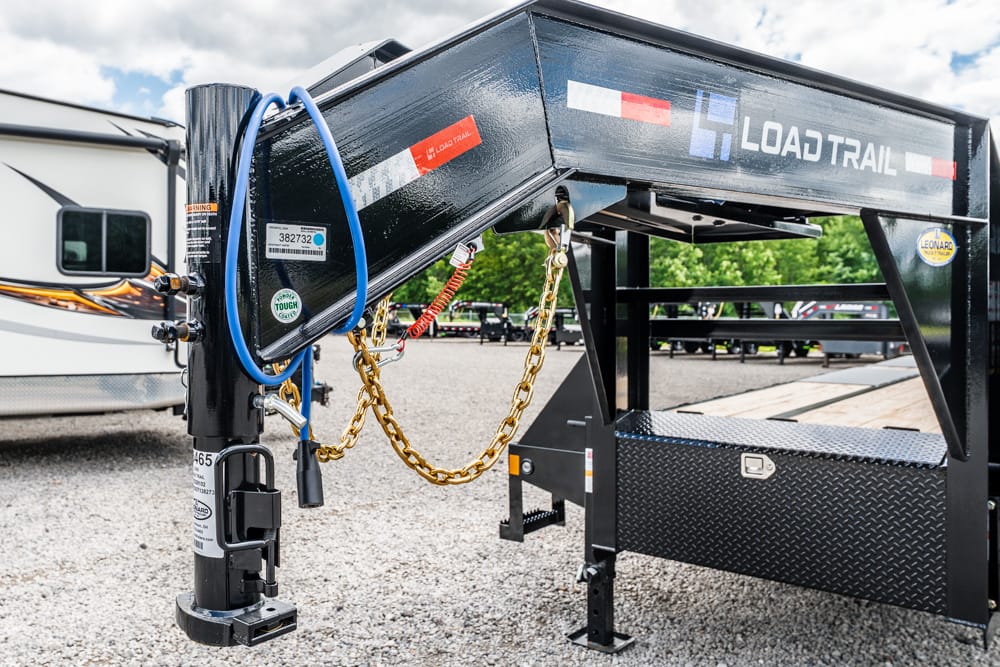 Close-up of a Load Trail gooseneck trailer hitch with safety chains, blue hose, and a jack on a gravel lot.