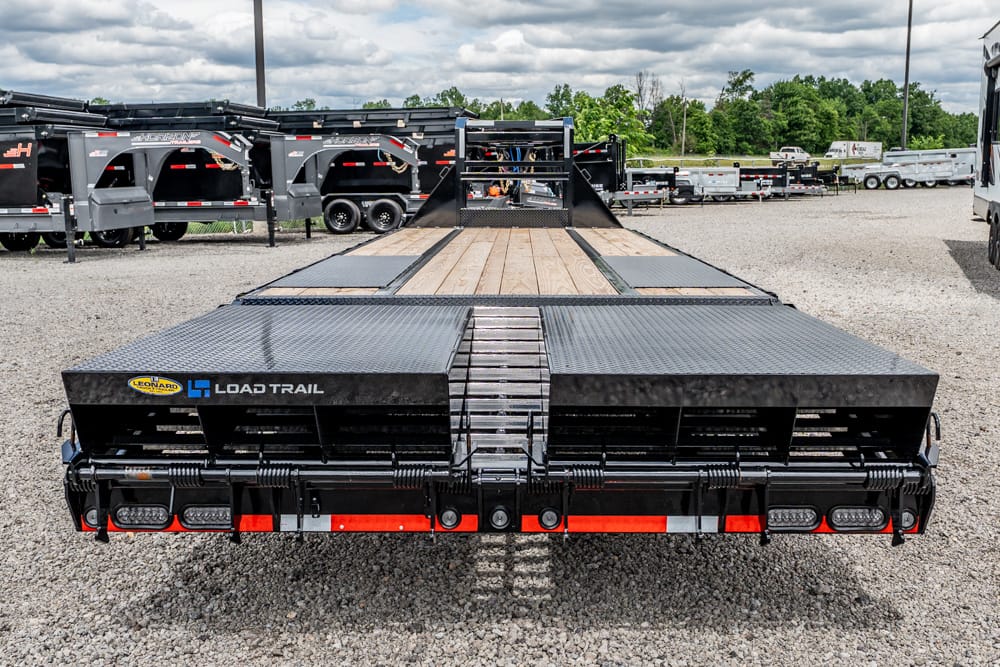 Rear view of a black Load Trail flatbed trailer with a wooden deck and diamond-plate ramps in a gravel trailer lot.