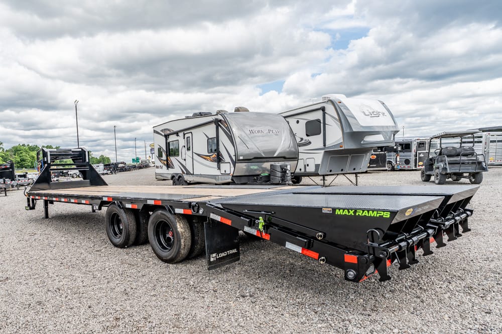 Heavy-duty flatbed trailer with MAX RAMPS in a RV lot, gravel ground and campers in the background.