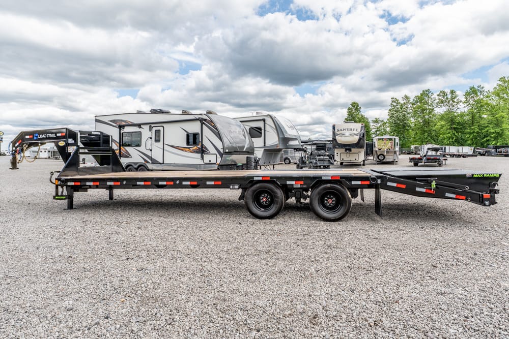 Heavy-duty flatbed trailer with tandem axles sits on a gravel lot, RVs and campers in the background ready for transport or storage.