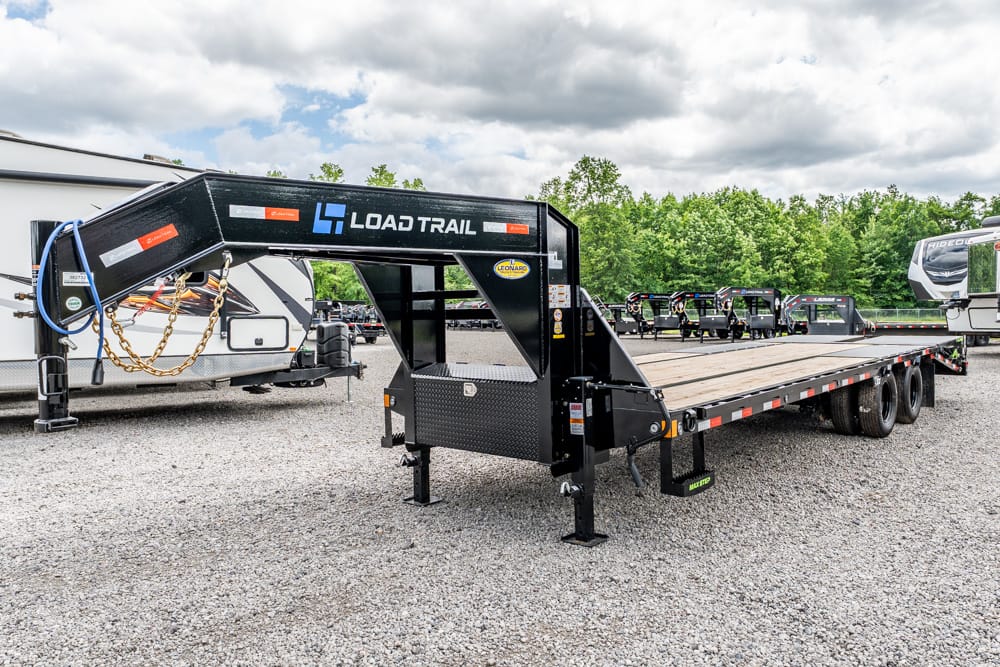 Black Load Trail gooseneck flatbed trailer with a wooden deck parked on a gravel lot, with RVs in the background.