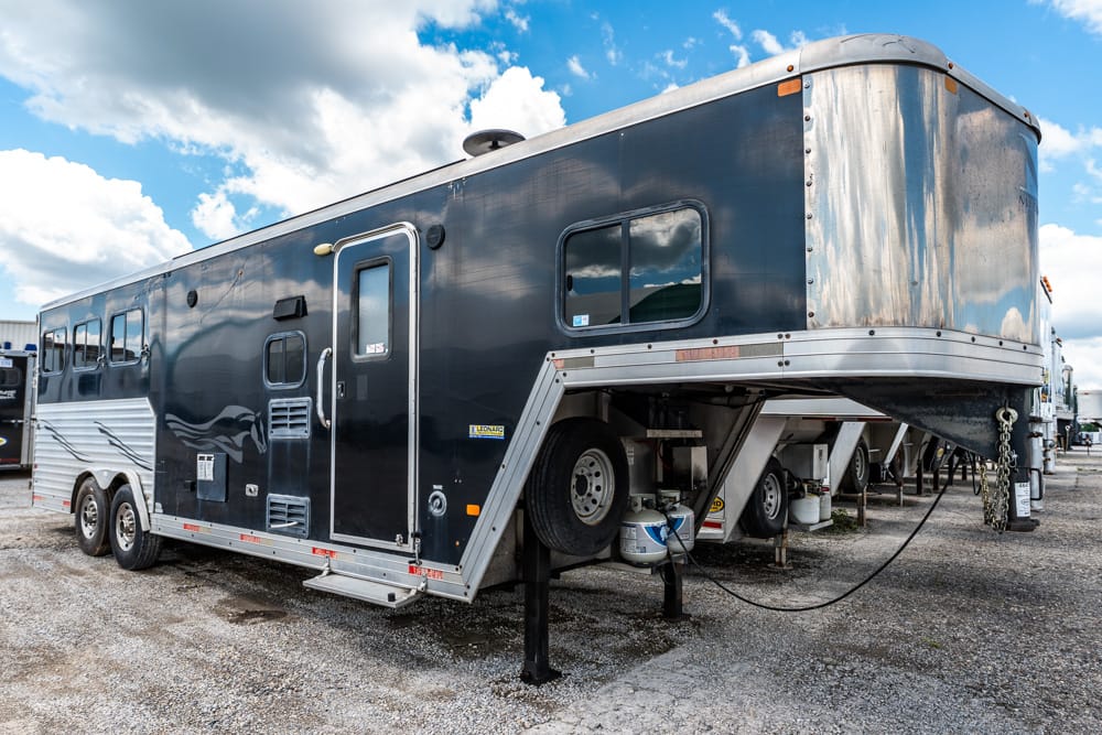 Large dark horse trailer with living quarters, raised hitch and stabilizing jacks in a gravel lot under a blue sky with clouds