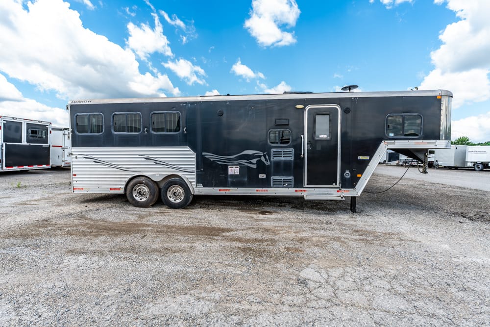 Black two-axle horse trailer with silver lower siding and gooseneck hitch parked on a gravel lot under a blue sky.