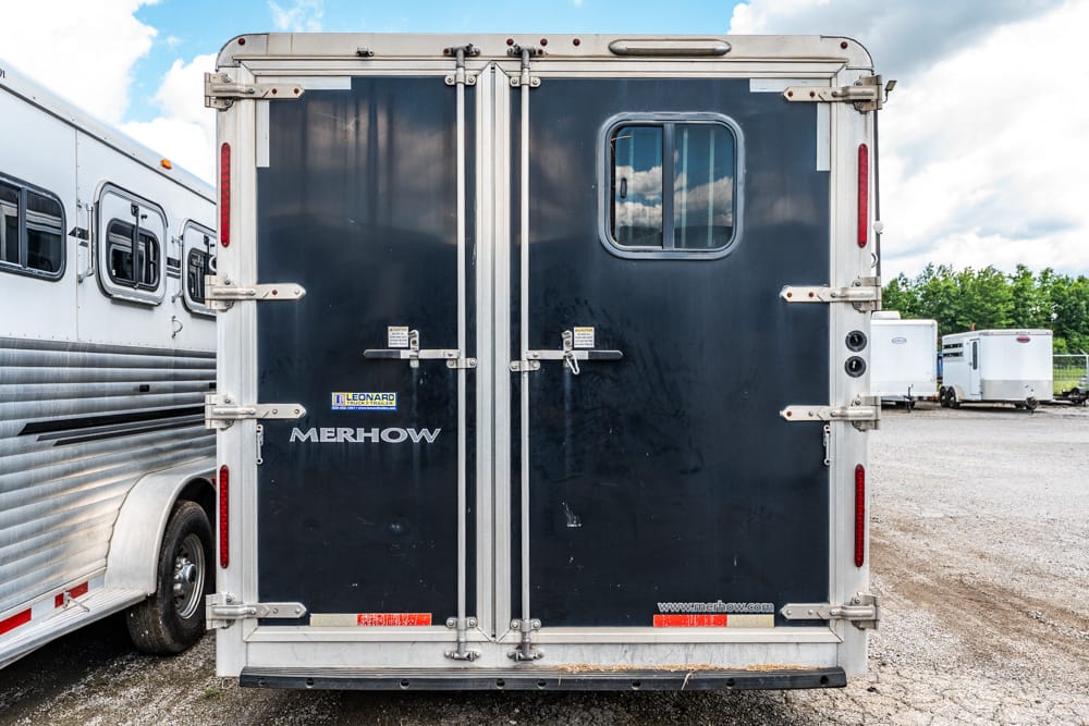 Rear view of a black Merhow horse trailer with double doors, silver hardware, and a small window on a gravel lot.