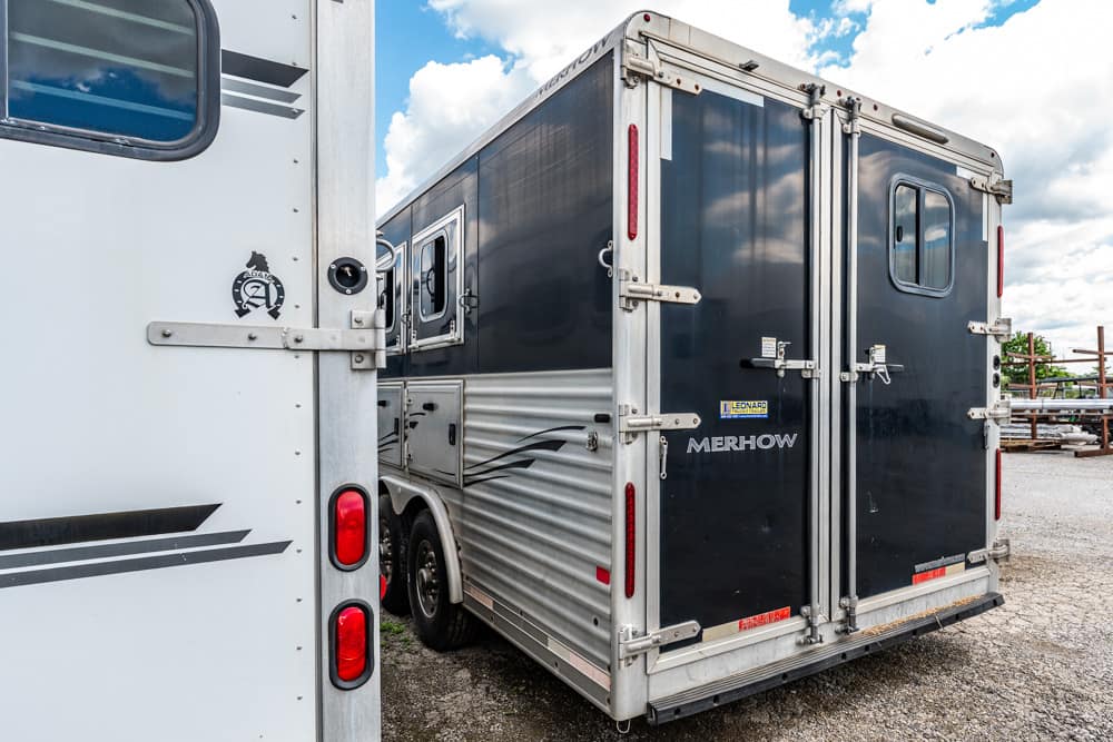 Rear view of a dark gray Merhow horse trailer with double doors and metal framing in a gravel lot.