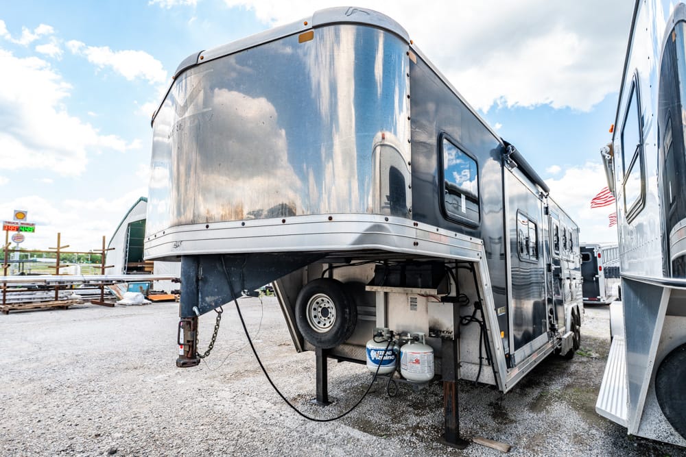 Shiny silver travel trailer parked in a gravel lot with a spare tire and propane tanks under the hitch visible beneath the front end