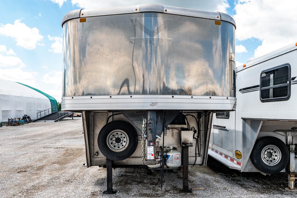 Front of a large aluminum trailer raised on its jacks in a gravel lot, with a spare tire and propane tank beneath.