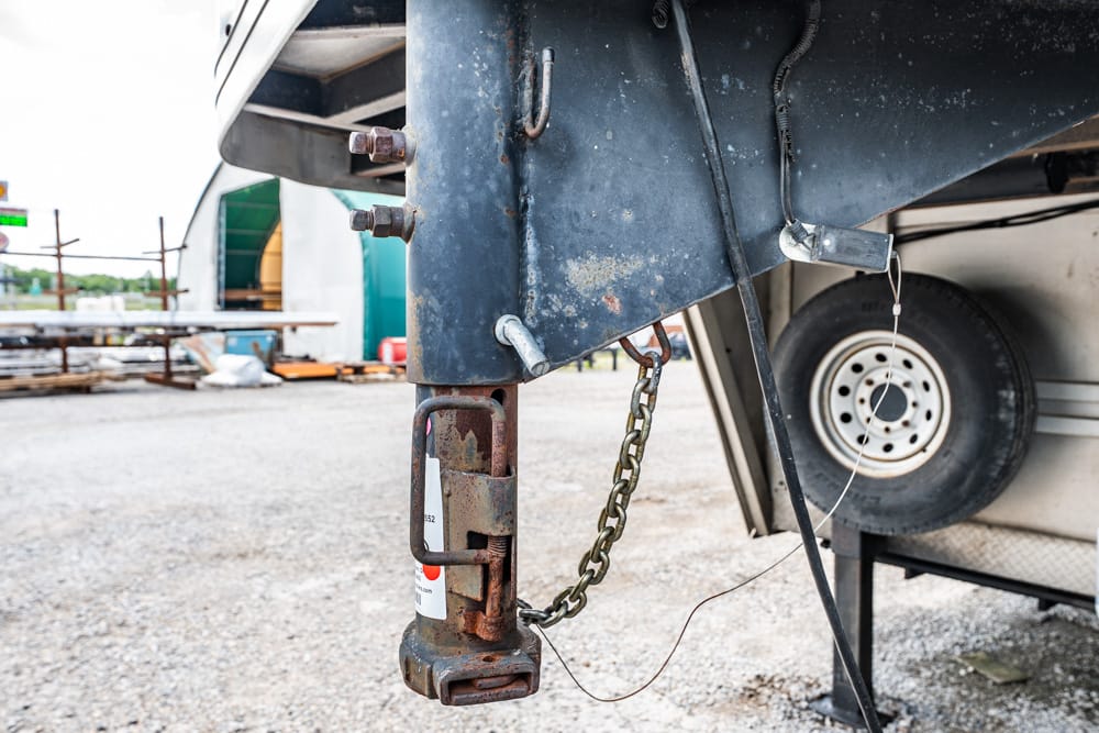 Rusty trailer hitch and jack with a chain, tire on a trailer in an outdoor gravel yard, casual industrial scene