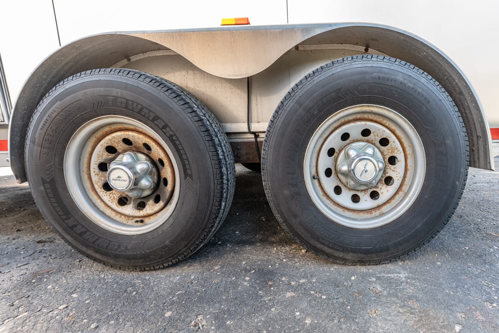Pair of dual trailer wheels with rusted steel rims under a beige fender on a dirt surface, showing tire treads and hub caps.