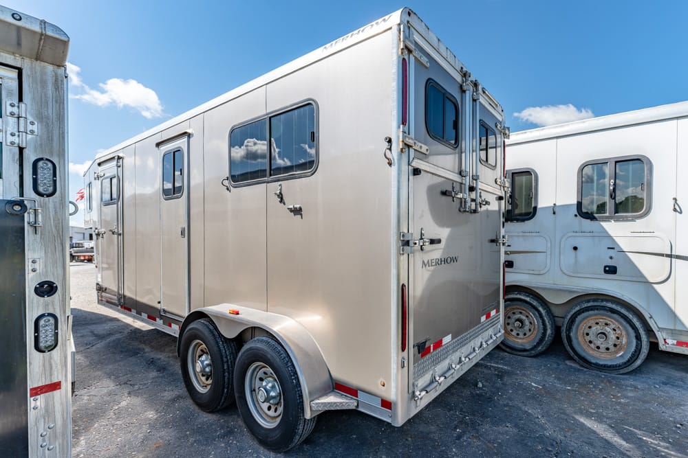 Silver Merhow horse trailer with tandem axles and side window parked on a gravel lot beside another trailer