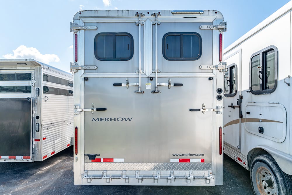 Rear view of a silver Merhow horse trailer with double doors, two small windows, and surrounding trailers on each side, under a blue sky with clouds.