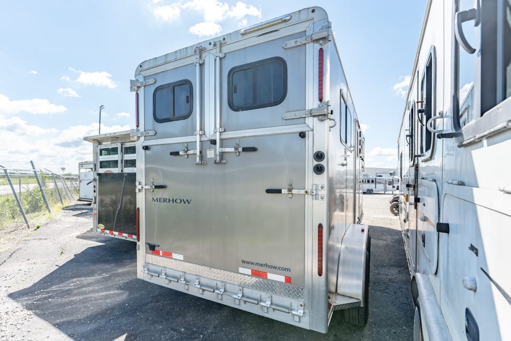 Rear view of a silver Merhow horse trailer with doors closed, parked between other trailers in a sunny lot:**