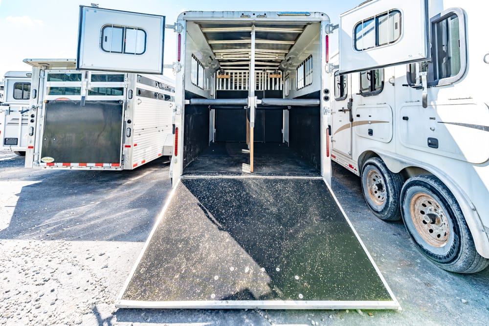 Open horse trailer with its ramp down, showing stalls and divider inside, parked between two other trailers.
