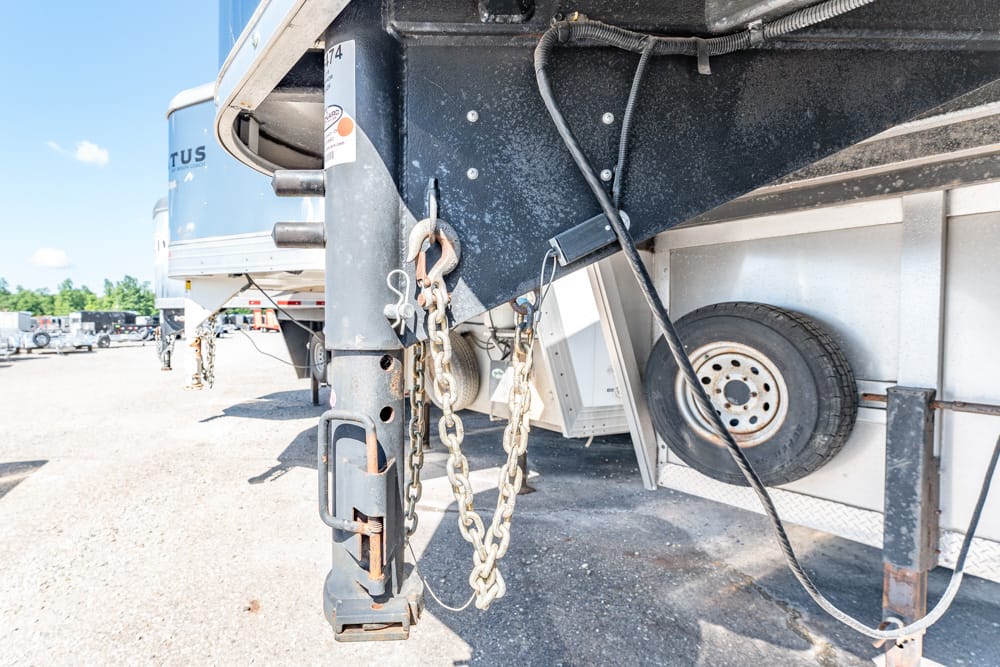 Close-up of a trailer hitch with rusty safety chains and a heavy electrical cable attached to a parked semi-trailer in a truck yard, under a sunny sky.